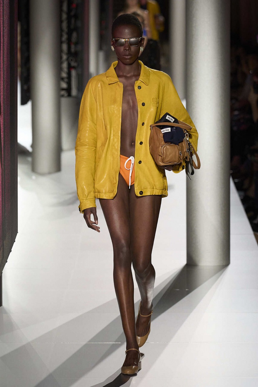 На фото один из образов коллекции весна-лето 2024 Miu Miu. Девушка идет в желтом долждевике и оранжевыми микро-шортами, а руках у нее большая сумка, на которую подвесили брелоки и шнурки. 